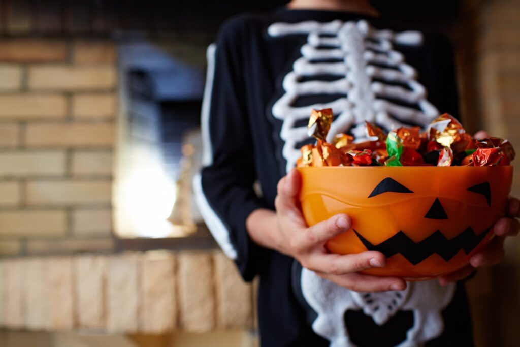 Child in a skeleton costume holding a jack-o'-lantern bowl of Halloween candy